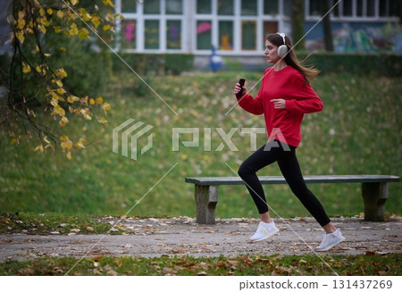 Young beautiful woman running in autumn park and listening to music with headphones on smartphone 131437269