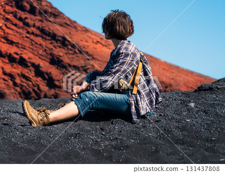 A girl sits on a black sand, enjoys a volcanic mountain landscape. 131437808