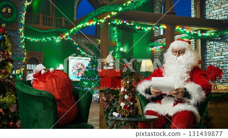 Man dressed as Santa Claus laughing, amused by funny letters received from kids. Jovial person in Santa costume entertained by comical wish lists written by children for Christmas, camera A 131439877