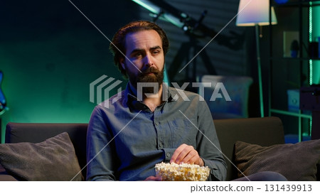 Sad man sitting on couch feeling heartbroken while watching tragic movie on streaming service. Depressed person seeing TV show favorite character dying, showing emotional discomfort, camera A 131439913