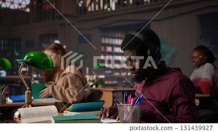 Black guy using study music to help with deep focus for academic success, consulting the campus university database to find reliable sources. Male college student fights for diploma. Camera B. 131439927