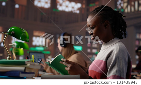 African american girl reading and taking notes for exam at the college, studying political science for upcoming class lesson in a library at university. Young student consults textbooks. Camera A. African american girl reading and taking notes for exam at the college, studying political science for upcoming class lesson in a library at university. Young student consults textbooks. Camera A. 131439945