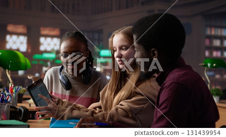 Multicultural students group discussing a financial project for a university course, using laptop and textbooks with class notes in the library. Entrepreneurship and management skills. Camera A. 131439954