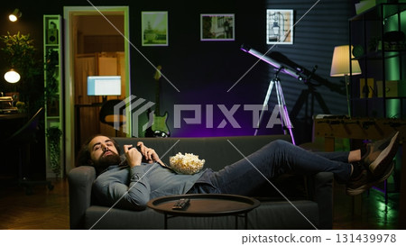 Gamer lounged on couch focused on winning competitive multiplayer game eating popcorn. Person in home theatre holding controller munching on salty snack, playing videogames, camera B Gamer lounged on couch focused on winning competitive multiplayer game eating popcorn. Person in home theatre holding controller munching on salty snack, playing videogames, camera B 131439978