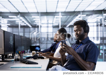 Server farm admin with headphones on texting friends on smartphone and using artificial intelligence tech. Data center worker using mobile phone, taking break from implementing AI 131440036