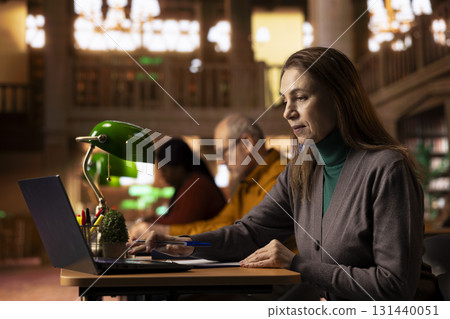 Adult learner transcribes information during a lecture in a university library, surrounded by academic textbooks and archived documents. Higher education personal goals on campus. 131440051