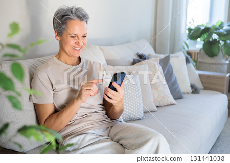 Happy senior woman using smartphone on sofa at home, staying connected and enjoying technology 131441038