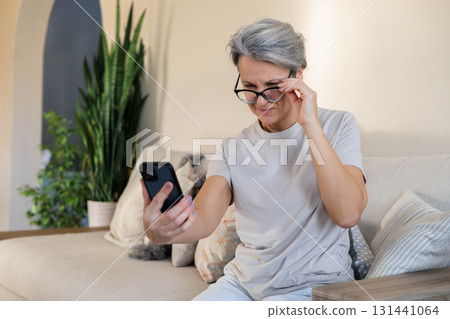 Senior woman in glasses using smartphone at home, squinting and adjusting vision Senior woman in glasses using smartphone at home, squinting and adjusting vision 131441064