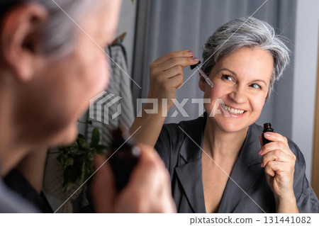 An adult beautiful woman of age during menopause with gray hair applies serum against wrinkles 131441082