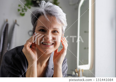 Mature happy beautiful woman in menopause with grey hair removing makeup from her face 131441084