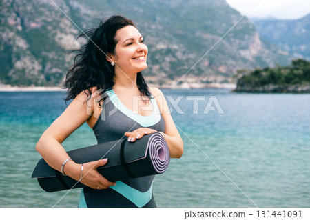 Smiling woman holding yoga mat by the sea, enjoying nature and healthy coastal lifestyle 131441091