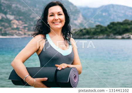 Happy woman holding yoga mat by the sea, enjoying outdoor fitness in peaceful nature 131441102