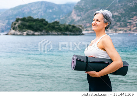 Happy senior woman with yoga mat on beach, embracing fitness and aging gracefully outdoors 131441109