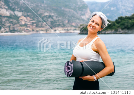 Smiling senior woman with yoga mat by the sea, embracing healthy aging and active lifestyle 131441113