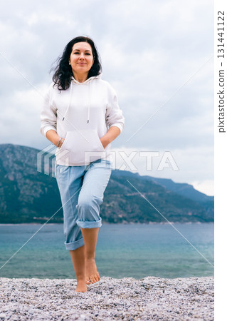 Confident Woman in Hoodie Standing Barefoot on Pebble Beach with Mountain and Sea Background 131441122