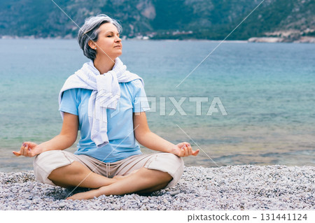 Mature woman meditating by the sea, enjoying peaceful moment on rocky beach with mountains Mature woman meditating by the sea, enjoying peaceful moment on rocky beach with mountains 131441124