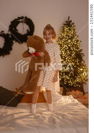 Girl hugging teddy bear with Santa hat front view sitting on bed near Christmas tree with lights Girl hugging teddy bear with Santa hat front view sitting on bed near Christmas tree with lights 131441590