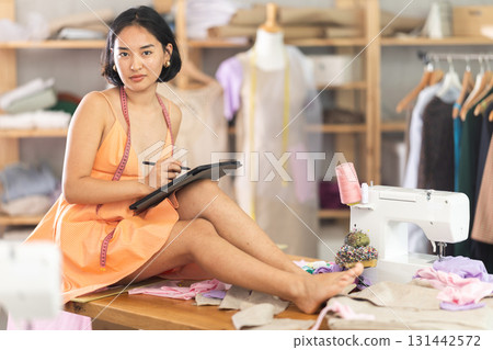 Seamstress girl draws outline of future dress on an electronic tablet in sewing workshop Seamstress girl draws outline of future dress on an electronic tablet in sewing workshop 131442572