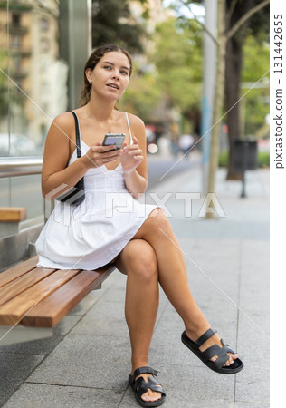 Positive young girl sitting at bus stop holding phone in hand 131442655