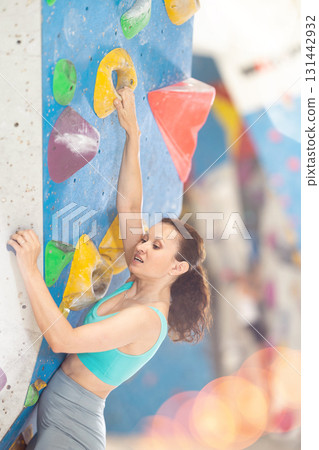 Woman in comfortable athletic clothing moving carefully on bouldering wall clinging to colorful hand and foot holds 131442932