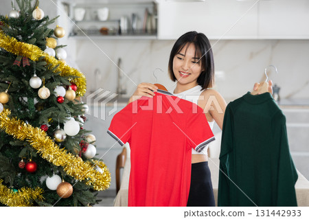 Happy young Asian girl trying to choose a dress standing next to Xmas-tree 131442933