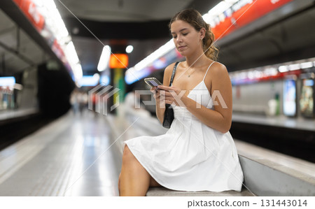 Positive young girl sitting on subway platform holding phone in hand Positive young girl sitting on subway platform holding phone in hand 131443014