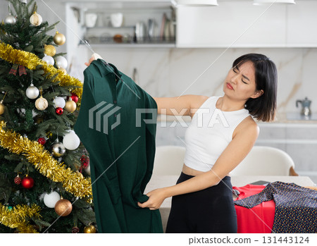 Sad young Asian girl trying to choose a dress standing next to Xmas tree 131443124
