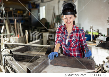 Portrait of a female welder standing with a welding semi-automatic machine and a safety helmet in metal machining workshop 131443132