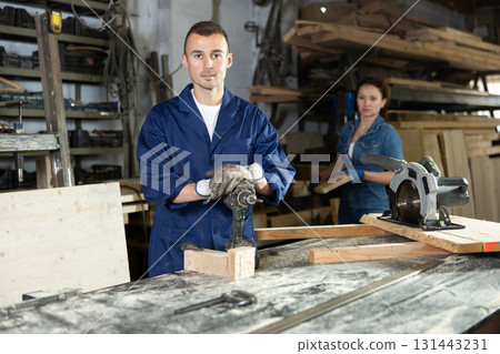 Male employee of carpentry workshop uses drill to make holes in wooden billet Male employee of carpentry workshop uses drill to make holes in wooden billet 131443231