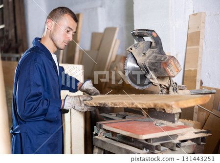 Male employee of workshop cuts wooden board into narrow parts using circular saw 131443251