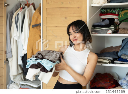 Happy young Asian girl holding some items of clothing by the wardrobe Happy young Asian girl holding some items of clothing by the wardrobe 131443269
