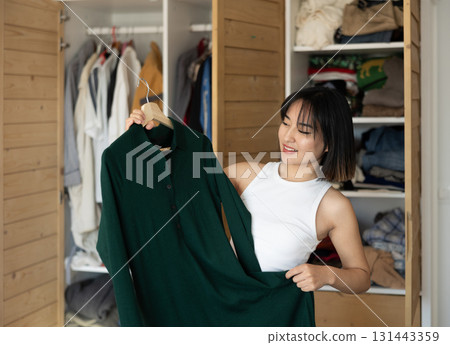 Happy young Asian girl trying to choose a dress standing by wardrobe 131443359