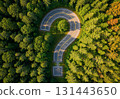 An aerial view shows a road forming a question mark surrounded by lush green forest. The unique winding path is embedded in dense trees, creating a striking visual contrast from above against the natu 131443650