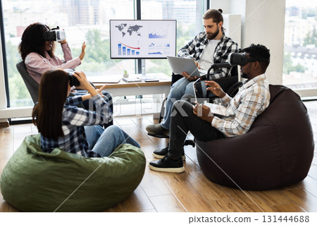 Group of diverse adults using virtual reality headsets and laptops in modern office setting. Individuals of various backgrounds collaborating in technology-driven workspace fostering creativity 131444688