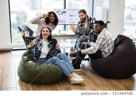 Young diverse coworkers relaxing on beanbags, taking selfie using smartphone, enjoying modern office. Multiracial group of adult friends casually interacting, inclusive team-building Young diverse coworkers relaxing on beanbags, taking selfie using smartphone, enjoying modern office. Multiracial group of adult friends casually interacting, inclusive team-building 131444788