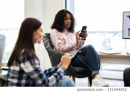 Two young adult women in casual clothing using digital devices in a bright office. One woman using smartphone while the other interacts with a tablet, representing communication, technology 131444852