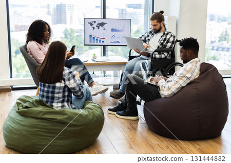 Group of young adults, diverse gender and race, working together in modern casual office. Collaborative atmosphere, bean bags, wheelchair accessibility, and digital devices convey teamwork, inclusion. Group of young adults, diverse gender and race, working together in modern casual office. Collaborative atmosphere, bean bags, wheelchair accessibility, and digital devices convey teamwork, inclusion. 131444882
