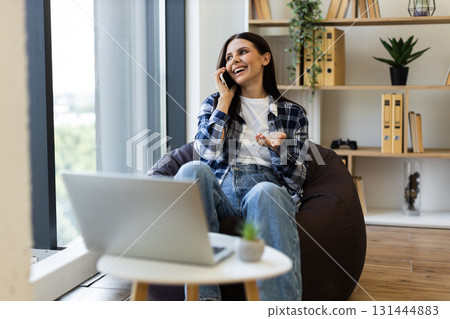 Smiling young adult Caucasian woman having phone conversation sitting on bean bag near window Smiling young adult Caucasian woman having phone conversation sitting on bean bag near window 131444883