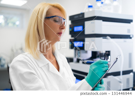 Middle aged blonde scientist analyzing data and writing notes in professional laboratory next to LC MS. Concept of quality research. Middle aged blonde scientist analyzing data and writing notes in professional laboratory next to LC MS. Concept of quality research. 131446944