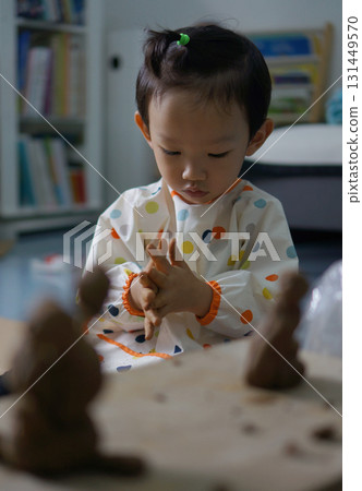 A young child focused on playing with clay, engaged in the process of creation. The child is seated at a wooden table, molding the clay with focus and care A young child focused on playing with clay, engaged in the process of creation. The child is seated at a wooden table, molding the clay with focus and care 131449570