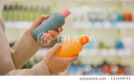 Close-up of female hands comparing cosmetic bottles in a beauty store. Woman reading label and checking ingredients while choosing haircare or body care product. Concept of conscious retail shopping. Close-up of female hands comparing cosmetic bottles in a beauty store. Woman reading label and checking ingredients while choosing haircare or body care product. Concept of conscious retail shopping. 131449741