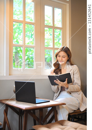 Confident businesswoman smiling while writing notes in a notebook beside a laptop 131452641
