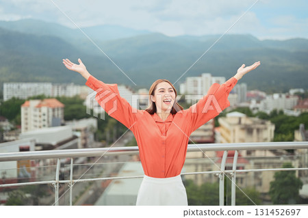 Cheerful woman enjoying freedom with arms open wide against urban skyline and mountains 131452697
