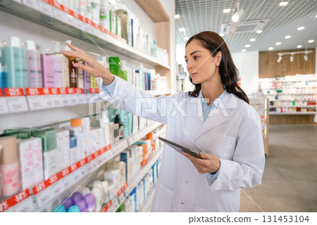 Female pharmacist with clipboard checking products on pharmacy shelves 131453104