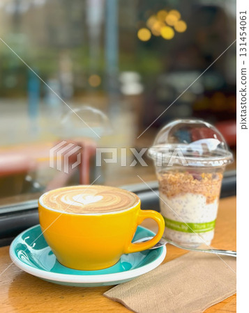 Yellow cup of cappuccino with latte art sits beside a granola yogurt parfait on a wooden table near a cafe window. Concept of coffee, breakfast, healthy lifestyle in a relaxed atmosphere 131454061