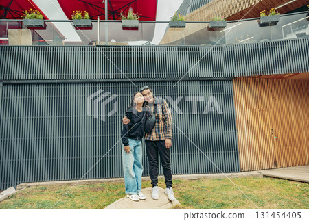 A young Asian woman and a young Hispanic man stand together, smiling in a recreational area with modern architecture and greenery. 131454405