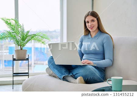 Portrait of young woman sitting on chair at home with laptop looking at camera Portrait of young woman sitting on chair at home with laptop looking at camera 131455212