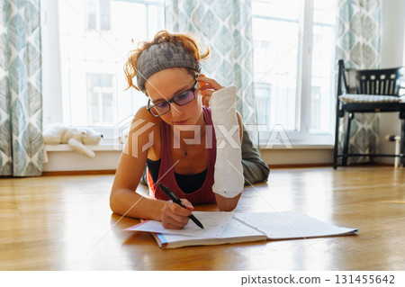 Teenage girl with broken arm in cast, wearing glasses, doing homework, at home 131455642