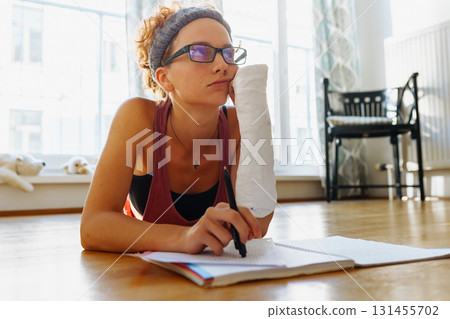 Teenage girl with broken arm in cast, wearing glasses, doing homework, at home 131455702