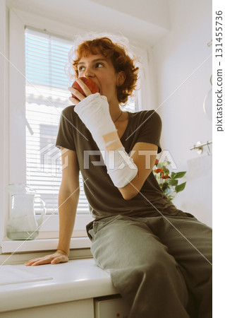 teenage girl with broken arm in cast in kitchen sitting on countertop eating apple 131455736